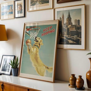May include: A framed print featuring a golden retriever playing basketball, with the text "KEEP HOOPING" in red. The dog is jumping to dunk the ball. The print is in a gold frame and is displayed with other framed art.