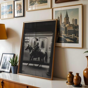 May include: A framed black and white photograph of a person riding a horse inside a subway car. The image is displayed with other framed art pieces, a lamp, and decorative vases on a white surface.