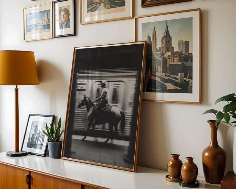 May include: A framed black and white photograph of a person riding a horse inside a subway car. The image is displayed with other framed art pieces, a lamp, and decorative vases on a white surface.