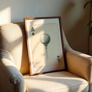 May include: A framed print of a golf ball with an ace of spades playing card design. The print is in a brown frame and is hanging on a beige wall.