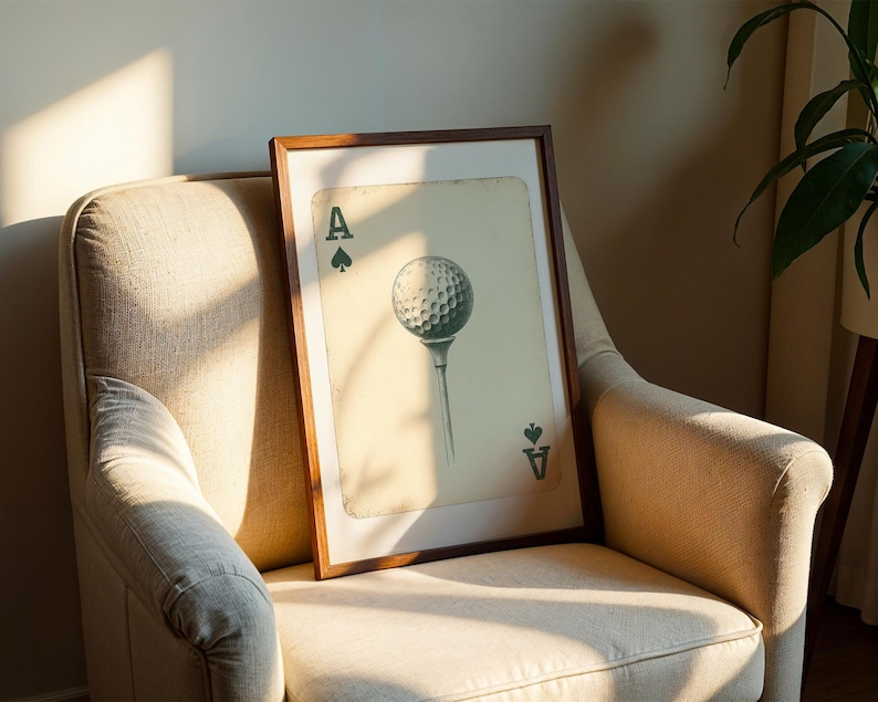 May include: A framed print of a golf ball with an ace of spades playing card design. The print is in a brown frame and is hanging on a beige wall.