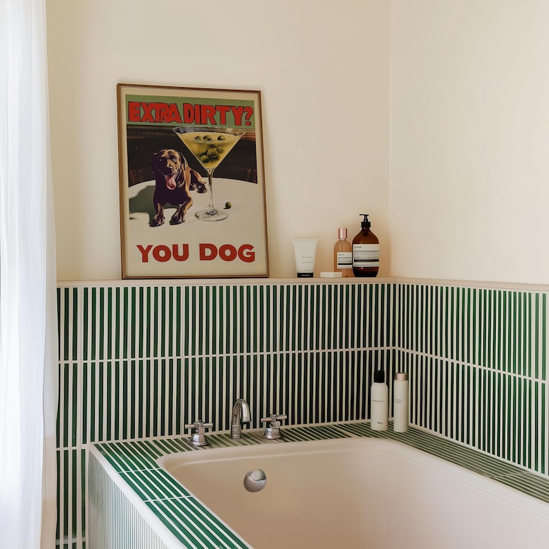 May include: A white bathtub with a green tiled surround. The tiles are arranged in vertical stripes. Above the tub is a shelf with a framed poster that says "Extra Dirty? You Dog" and several bottles of toiletries.