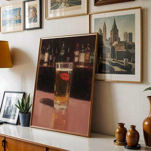 Puede incluir: Una impresi&oacute;n enmarcada de un vaso de cerveza con una huella de labio rojo, exhibida sobre una superficie blanca. La obra de arte forma parte de una pared de galer&iacute;a con otras impresiones enmarcadas y una l&aacute;mpara. Tambi&eacute;n se ven un mueble de madera y jarrones decorativos.