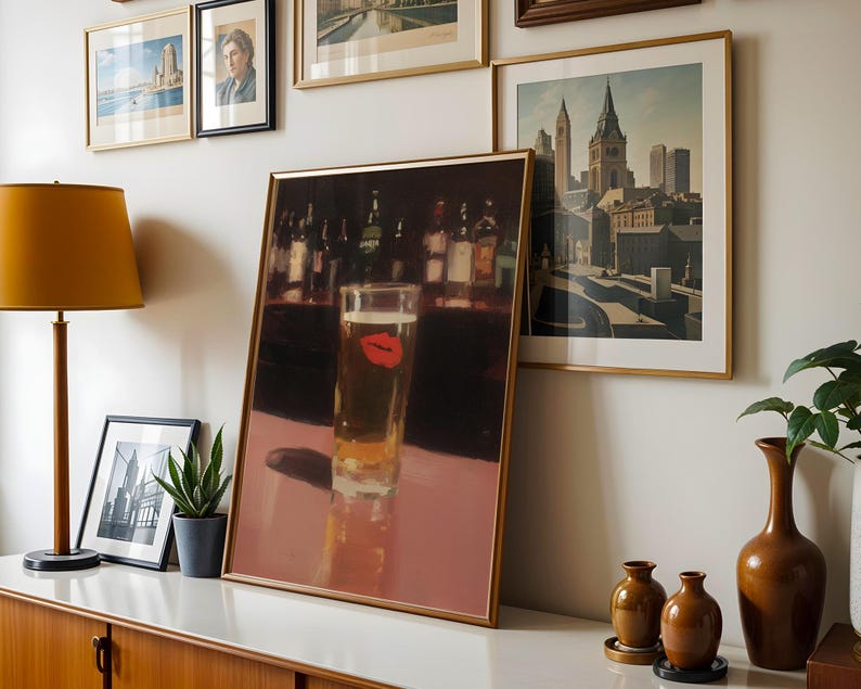 Puede incluir: Una impresi&oacute;n enmarcada de un vaso de cerveza con una huella de labio rojo, exhibida sobre una superficie blanca. La obra de arte forma parte de una pared de galer&iacute;a con otras impresiones enmarcadas y una l&aacute;mpara. Tambi&eacute;n se ven un mueble de madera y jarrones decorativos.