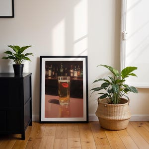 Puede incluir: Una impresi&oacute;n enmarcada que muestra un vaso de cerveza con una marca de l&aacute;piz labial rojo. La impresi&oacute;n est&aacute; en un marco negro, sobre un suelo de madera. Un armario negro y plantas en macetas tambi&eacute;n est&aacute;n en la habitaci&oacute;n.