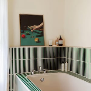 May include: A framed print depicts a martini glass and billiard balls on a green pool table. The bathroom features a white bathtub, green and white striped tile, and toiletries on a shelf.