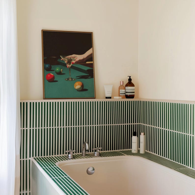 May include: A framed print depicts a martini glass and billiard balls on a green pool table. The bathroom features a white bathtub, green and white striped tile, and toiletries on a shelf.