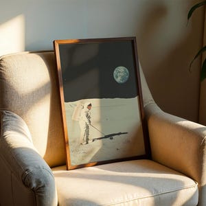 May include: A framed print of an astronaut in a spacesuit walking on the moon, with a small planet in the background. The print is in a brown wooden frame and is hanging on a beige wall.