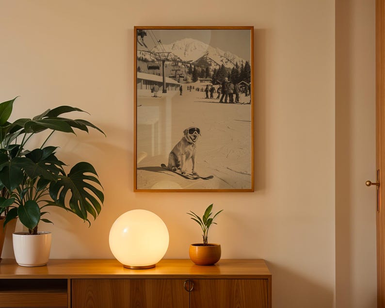 May include: A framed black and white print of a dog wearing sunglasses on skis, set against a snowy mountain backdrop. The print is in a wooden frame, hanging above a wooden cabinet with a glowing orb lamp and potted plants.