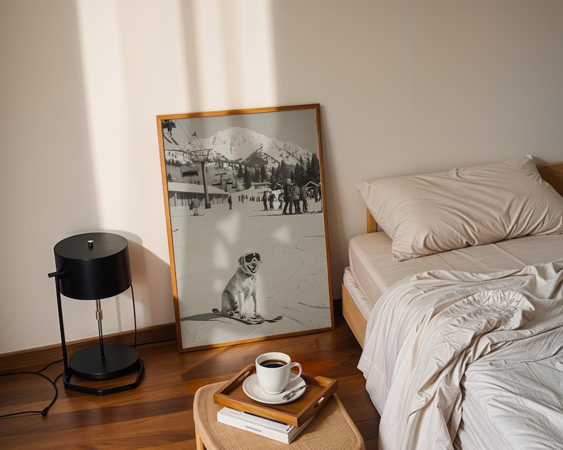 May include: A framed black and white photograph of a dog wearing goggles on skis, set against a snowy mountain backdrop. A black lamp, a wooden tray with a coffee cup, and a bed with beige bedding are in the foreground.