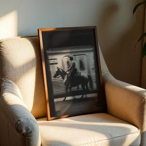 May include: A framed black and white photograph of a cowboy riding a horse in front of a subway train. The image is displayed in a dark wooden frame and is resting on a beige armchair. The scene is illuminated by natural light.