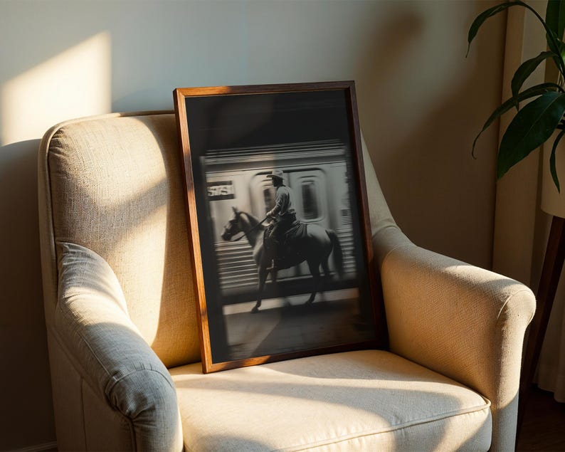 May include: A framed black and white photograph of a cowboy riding a horse in front of a subway train. The image is displayed in a dark wooden frame and is resting on a beige armchair. The scene is illuminated by natural light.