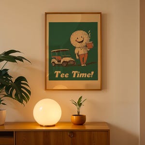 May include: A framed poster with a green background featuring a cartoon golf ball character holding a cup of coffee. The character is standing next to a golf cart. The poster has the text "Tee Time!" printed on it. The poster is hanging on a wall above a wooden table with a white lamp and a potted plant.