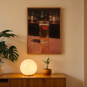 Puede incluir: Una obra de arte enmarcada representa un vaso de cerveza con una huella de labio rojo, sobre un fondo de bar. Una l&aacute;mpara esf&eacute;rica brillante se encuentra sobre un mueble de madera, con una planta en maceta a la derecha y una planta m&aacute;s grande a la izquierda.