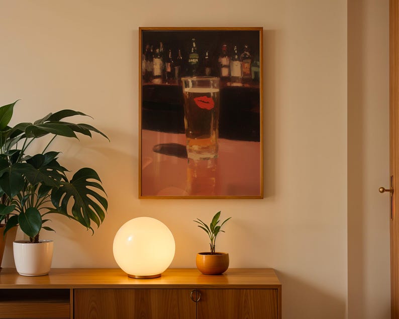 Puede incluir: Una obra de arte enmarcada representa un vaso de cerveza con una huella de labio rojo, sobre un fondo de bar. Una l&aacute;mpara esf&eacute;rica brillante se encuentra sobre un mueble de madera, con una planta en maceta a la derecha y una planta m&aacute;s grande a la izquierda.