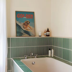 May include: A bathroom scene featuring a framed poster with the words "GOLDEN HOUR" above a dog surfing. The room has green and white striped tile, a white bathtub, and toiletries on a shelf.