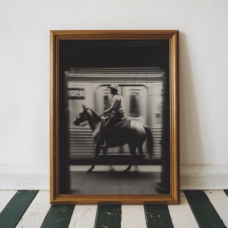 May include: Framed black and white photograph of a cowboy riding a horse in front of a speeding subway train. The image is in a gold-colored wooden frame, set against a white wall and a black and white striped floor.