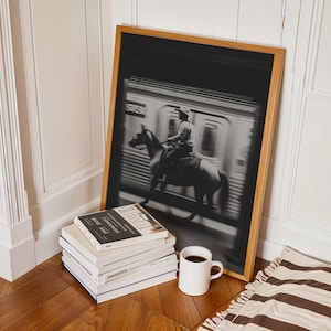May include: A framed black and white photograph depicts a person on horseback riding past a subway train. The wooden frame leans against a wall, next to a stack of books and a white mug filled with coffee. A striped rug is on the floor.