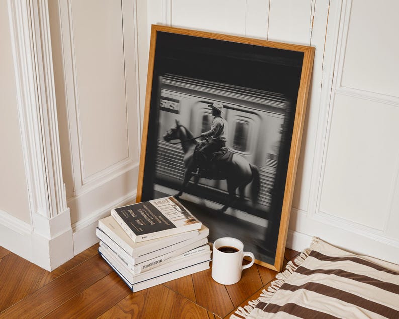 May include: A framed black and white photograph depicts a person on horseback riding past a subway train. The wooden frame leans against a wall, next to a stack of books and a white mug filled with coffee. A striped rug is on the floor.