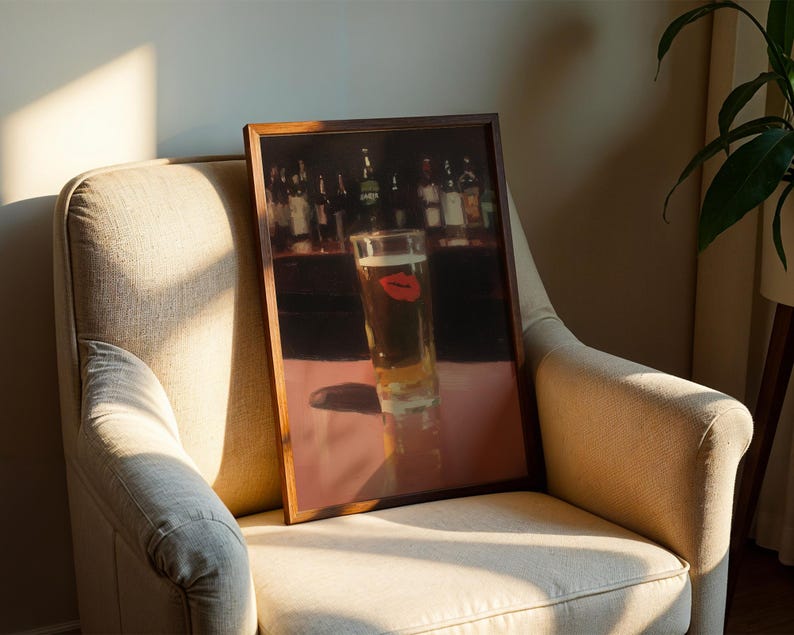 Puede incluir: Una impresi&oacute;n enmarcada de un vaso de cerveza con una huella de labio rojo, sobre un fondo de bar. La obra de arte se exhibe en un sill&oacute;n beige, con sombras de la luz solar.
