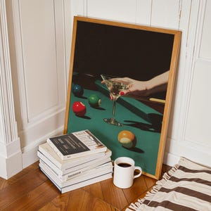 May include: A framed print featuring a hand holding a martini glass over a pool table with billiard balls. A stack of books, a white mug with coffee, and a striped rug are in the foreground. The frame is made of light brown wood.