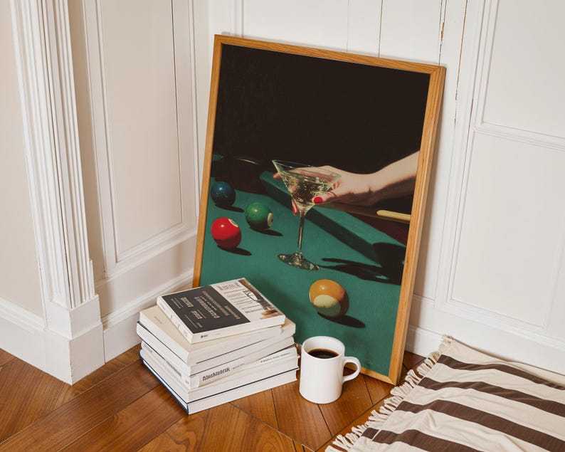 May include: A framed print featuring a hand holding a martini glass over a pool table with billiard balls. A stack of books, a white mug with coffee, and a striped rug are in the foreground. The frame is made of light brown wood.