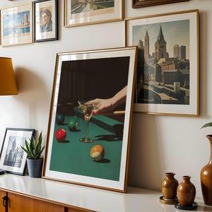 May include: A framed print of a martini glass on a green pool table, with a hand reaching for the glass. The image is surrounded by other framed art and decor, including a lamp and vases. The print has a gold frame.