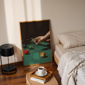 May include: A framed print depicts a hand holding a martini glass over a pool table with colorful billiard balls. A black lamp, a bed with beige bedding, and a coffee cup on a tray are also visible.
