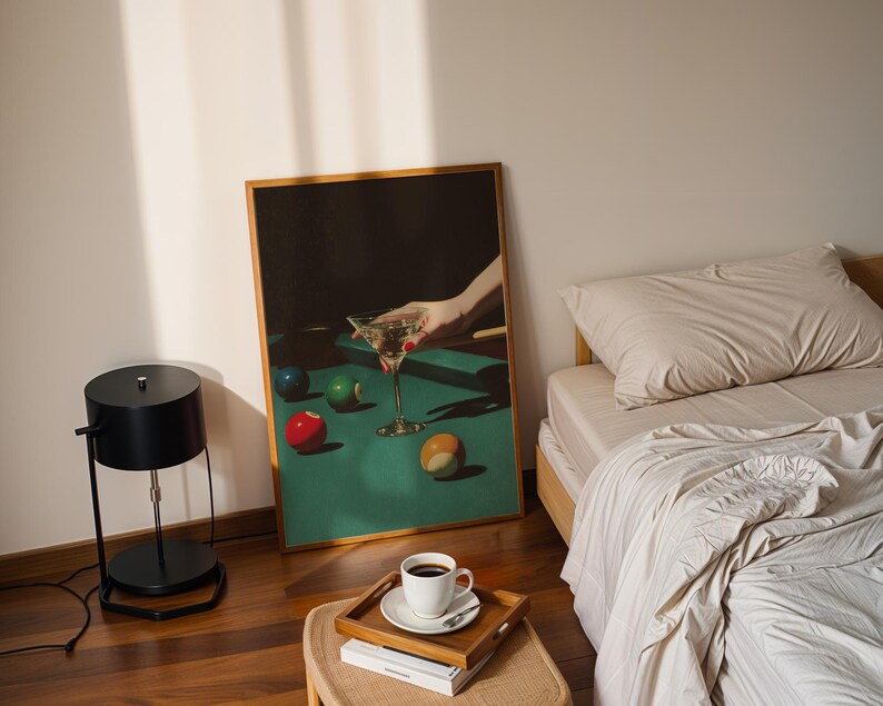 May include: A framed print depicts a hand holding a martini glass over a pool table with colorful billiard balls. A black lamp, a bed with beige bedding, and a coffee cup on a tray are also visible.