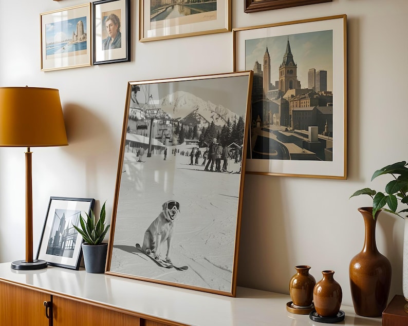 May include: A framed black and white photograph of a dog wearing goggles on skis, with a snowy mountain backdrop. The image is displayed on a white surface, alongside other framed art and a lamp with a mustard-yellow shade. A plant in a gray pot is also present.