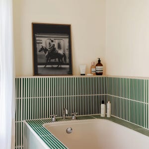 May include: A bathroom scene featuring a framed black and white photograph of a person riding a horse. The room has green and white striped tile, a white bathtub, and toiletries on a shelf. A white curtain is visible.