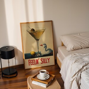 Puede incluir: Un p&oacute;ster de estilo vintage con una copa de martini y un pato, con el texto "Feelin' Silly". El p&oacute;ster est&aacute; enmarcado en madera y est&aacute; colgado en una pared blanca. Tambi&eacute;n se puede ver una l&aacute;mpara negra y una cama con ropa de cama blanca en la imagen.