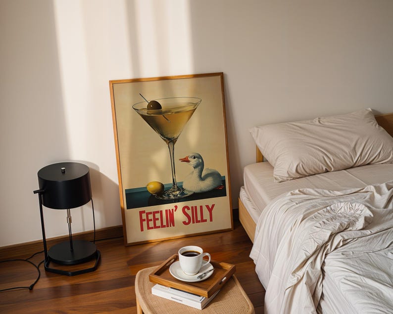 Puede incluir: Un p&oacute;ster de estilo vintage con una copa de martini y un pato, con el texto "Feelin' Silly". El p&oacute;ster est&aacute; enmarcado en madera y est&aacute; colgado en una pared blanca. Tambi&eacute;n se puede ver una l&aacute;mpara negra y una cama con ropa de cama blanca en la imagen.