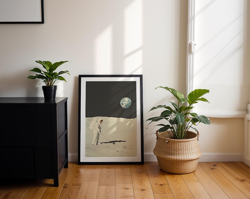 May include: A framed print of an astronaut playing golf on the moon with Earth in the background. The print is in a black frame and is hanging on a white wall.
