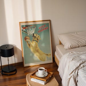 May include: A framed poster with a golden retriever dog playing basketball, with the words "KEEP HOOPING" in red. The poster is in a bedroom setting, with a bed, a side table, and a black lamp.