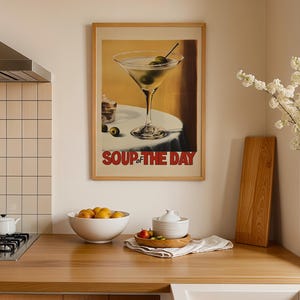 Peut inclure: Une affiche encadr&eacute;e avec un fond jaune repr&eacute;sentant un verre &agrave; martini avec des olives et le texte "Soup of the Day". L'affiche est accroch&eacute;e &agrave; un mur blanc au-dessus d'un comptoir en bois.