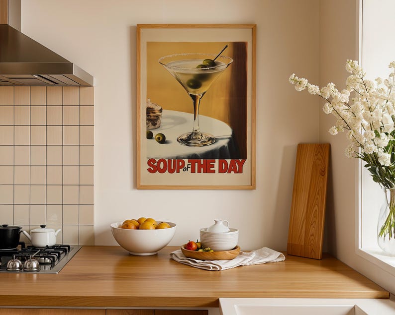 Peut inclure: Une affiche encadr&eacute;e avec un fond jaune repr&eacute;sentant un verre &agrave; martini avec des olives et le texte "Soup of the Day". L'affiche est accroch&eacute;e &agrave; un mur blanc au-dessus d'un comptoir en bois.