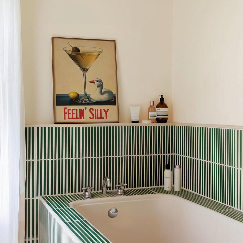 Puede incluir: Una ba&ntilde;era blanca con un borde de azulejos verdes. Los azulejos est&aacute;n dispuestos en rayas verticales. Sobre la ba&ntilde;era hay un estante con un cartel enmarcado que dice "Feelin' Silly" y una copa de martini con una aceituna y un pato. Tambi&eacute;n hay tres botellas de art&iacute;culos de tocador en el estante.