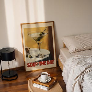 Peut inclure: Une affiche encadr&eacute;e avec un fond jaune et un verre &agrave; martini avec des olives. L'affiche dit "Soup of the Day" en lettres rouges. Une lampe noire avec un abat-jour rond est sur un sol en bois. Un lit avec une literie blanche est &agrave; l'arri&egrave;re-plan.