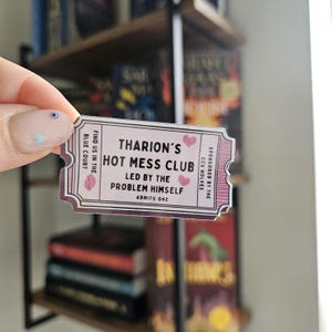 Op de afbeelding: Een rechthoekige sticker met een roze en wit ticketontwerp. De tekst op de sticker luidt "Tharion's Hot Mess Club" en "Led by the Problem Himself". De sticker bevat ook de tekst "Admits One" en "Find us in the Blue Court".