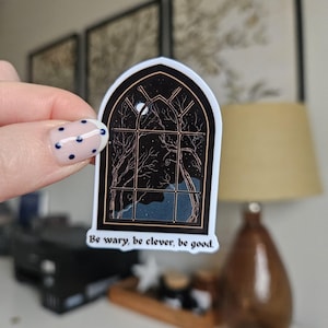 Può includere: Un adesivo con un design di finestra gotica con una scena di cielo notturno, tra cui una luna e alberi spogli. L'adesivo ha un bordo nero e il testo "Be wary, be clever, be good" in basso.