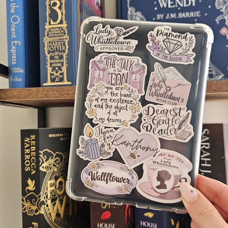 K&ouml;nnte beinhalten: Transparente Handyh&uuml;lle mit verschiedenen Aufklebern mit Texten und Illustrationen. Aufkleber enthalten S&auml;tze wie "Lady Whistledown Approved" und "Dearest Gentle Reader". Die H&uuml;lle wird in der N&auml;he eines B&uuml;cherregals mit B&uuml;chern gehalten.