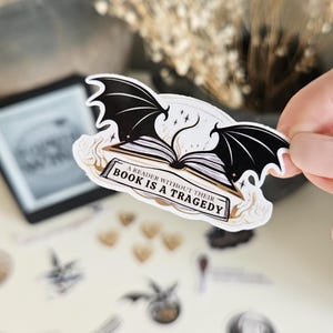 Peut inclure: Un autocollant représentant un livre avec des ailes de chauve-souris noires et le texte "A Reader Without Their Book Is A Tragedy". L'autocollant a une bordure blanche et un schéma de couleurs or et noir.