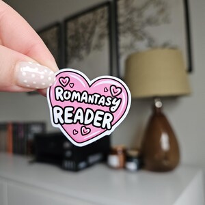 Op de afbeelding: Een roze hartvormige sticker met de woorden "Romantasy Reader" in het wit, omlijnd met zwart. De sticker heeft een roze rand en kleine hartillustraties. De sticker wordt door een hand omhoog gehouden.