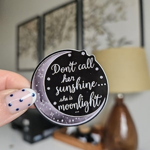 Può includere: Un adesivo rotondo nero con un design a mezzaluna e la citazione "Don't call her sunshine... she is moonlight" in scrittura bianca. L'adesivo ha un bordo bianco e dettagli a stella. L'adesivo è tenuto da una persona con una manicure.