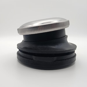 May include: A black rubber and metal fuel tank cap. The cap has a silver, ridged top and a black, layered rubber base. The cap is designed to seal a fuel tank.
