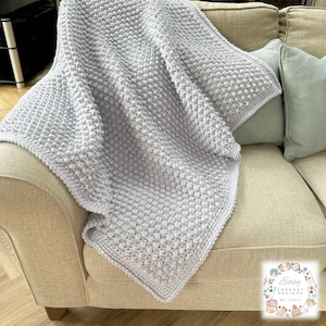 Peut inclure: Une couverture en crochet gris clair, avec un motif texturé en points en relief, drapée sur un canapé beige. Le motif de la couverture crée une sensation douce et tactile. Deux coussins bleu clair sont visibles. La couverture semble faite à la main.