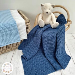 Può includere: Una copertina per neonati in crochet blu con un bordo in crochet bianco. Un orsacchiotto di peluche bianco è seduto sulla copertina.