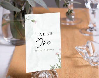 May include: A white table number card with green watercolor background and pink flowers. The card says "TABLE One" and "EMILY & DEAN". The card is on a clear glass stand.
