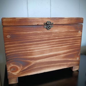 May include: A wooden box with a hinged lid and a brass latch. The box is made of natural wood with a reddish-brown stain. The box has a simple design and is perfect for storing small items.